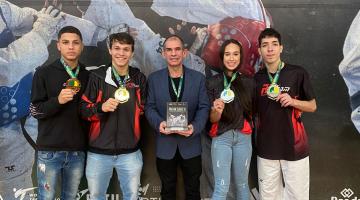 Treinador e atletas exibem medalhas e troféu em frente a banner do evento. #paratodosverem