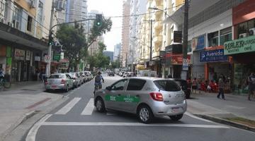 táxi está passando em rua sobre faixa de pedestres. Ao lado, há estabelecimentos comerciais. #paratodosverem