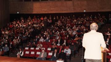 Auditório do Teatro Municipal com lotação quase máxima. No palco, de costas, um homem está à frente de uma tribuna falando ao público. #paratodosverem