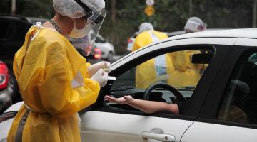 Mulher paramentada com avental, luva, gorro e máscaras faz teste em pessoa dentro de carro. #Paratodosverem