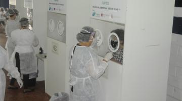 pessoas estão passando por teste nasal de covid atrás de biombos. Técnicos vestidos de branco procedem o exame do lado externo. #paratodosverem