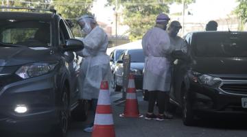 Carros estão parados em fila dupla. Pessoas de branco estão próximas às janelas fazendo os testes. #paratodosverem