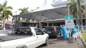 Fila de carros em estacionamento. Veículos se aproximam de uma tenda. Ao lado direito há uma faixa onde se lê Teste Rápido. Há pessoas usando aventais, máscaras e luvas para atender ao público que está nos carros. #Paratodosverem