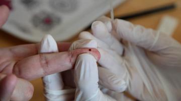 mãos com luvas seguram o dedo de uma pessoa que tem uma gota de sangue. #paratodosverem