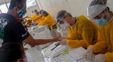 Várias pessoas paramentadas com avental, máscara, gorro e luva estão sentadas atrás de meses trabalhando na testagem de covid. Um homem estica o braço para ter retirada a gota de sangue. #Paratodosverem