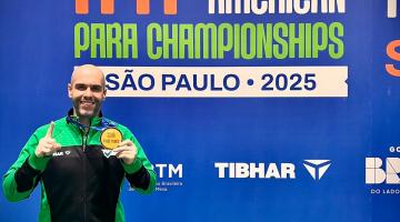 Mesatenista em pé junto ao painel do evento de tênis de mesa. #Paratodosverem