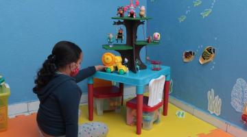 meninas está mexendo em brinquedo infantil em uma sala com piso emborrachado. Ela está de costas para a foto. #paratodosverem