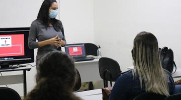 jovens estão sentados em sala de aula. Mulher está à frente de todos falando ao grupo. Há dois computadores atrás dela. - #paratodosverem