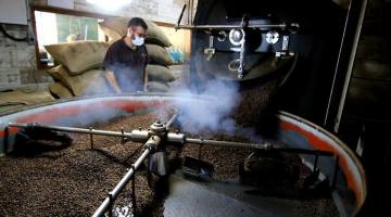 Equipamento de torra de café com diversos grãos. Máquina está em movimento. Homem está ao fundo diante de sacas de café empilhadas. #paratodosverem
