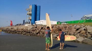 Surfistas se preparam para entrar no mar, com torre de jurados ao fundo. #pracegover.