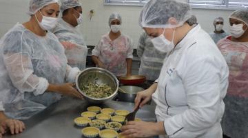 mulheres em primeiro plano coloca recheio em forminhas de torta sobre uma mesa. Outra mulher segura uma cumbuca com recheio. #paratodosverem