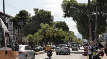 veículos trafegando em pista. Pessoas paradas em frente a uma faixa de pedestres aguardando para a travessia. #paratodosverem