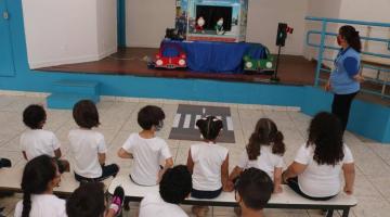 Mulher fala com crianças com teatro de fantoches ao fundo #paratodosverem