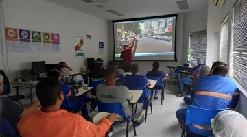 Profissionais sentados em uma sala observam explicações de homem apontando para telão #paratodosverem