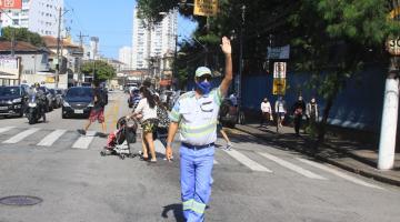 agente da CET está na rua, à frente de uma faixa de pedestres. Ele está com o braço esquerdo para o alto, fazendo gesto para veículos de 'pare'. Ao fundo, pessoas atravessam a rua. #paratodosverem