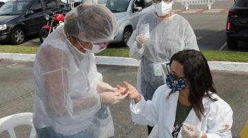 Duas mulheres estão em pé ao lado de uma sentada. Todas vestem avental branco, máscaras e luvas. Uma das que está de pé fura um dedo da mão direita da que está sentada. Uma outra está atrás acompanhando. Trata-se de um treinamento. #Paratodosverem