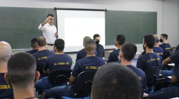 Homem está em pé, ao lado de telão, com a mão direita erguida ao falar com os guardas que estão sentados. #paratodosverem