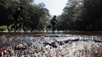 duas bicicletas passando sobre pequeno córrego em pedras com vegetação ao fundo