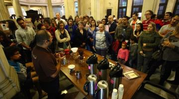 Várias pessoas assistem à preparação de café. Homem que está demonstrando fica atrás de mesa onde estão várias garrafas térmicas, coadores e copinhos de café. #paratodosverem