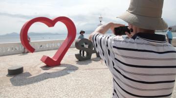 turista usando chapéu (de costas para a foto) usa celular para fotografar pessoa que posa atrás de monumento amo santos, diante da mureta da ponta da praia. #paratodosverem