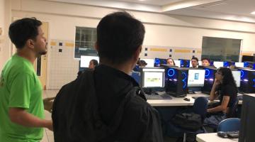 duas pessoas em pé e alunos sentados na frente de computadores #paratodosverem