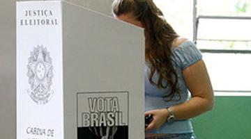 mulher vota atrás de cabine. Na cabine se justiça eleitoral, o brasão das armas nacional e cabine de votação. Na lateral, se lê Vota Brasil. Não se vê o rosto da mulher na cabine. #paratodosverem