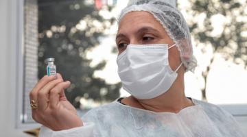 mulher usando avental, touca e máscara segura frasco com dose de vacina na mão direita. Ela olha o frasco.#paratodosverem