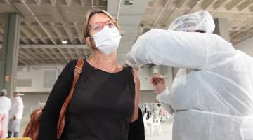 mulher está sendo vacinada em pé. Outra mulher, usando avental aplica a vacina. #paratodosverem