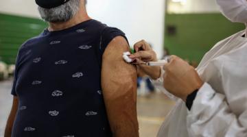 imagem em close de uma pessoa vacinando o braço de um homem, cuja camiseta está com a manga recolhida ao ombro. Ele usa máscara. #paratodosverem