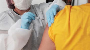 Mulher usando máscara, gorro, avental e luvas vacina uma pessoa que de costas, em close. #Paratodosverem