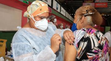 Idosa está de costas sendo vacinada por uma mulher no braço esquerdo. #Paratodosverem
