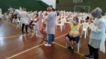 Ginásio de esportes com cadeiras enfileiradas. Na primeira filha há alguns idosos sentados aguardando a vacinação. Os aplicadores estão em pé. #paratodosverem