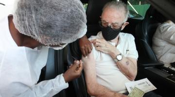 Mulher vestida de branco vacina idoso dentro de carro. Ele está segurando a manga da blusa e tem documentos no colo. #Paratodosverem