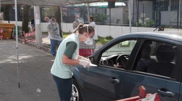 Mulher usando máscara está falando com motorista dentro de carro. À frente deles está armada uma base com cobertura onde se lê Samu Prefeitura de Santos
