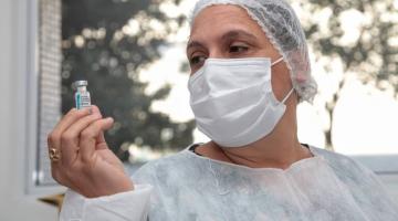 mulher com avental branco, mascara e touca olha para frasco de vacina que está na sua mão direita. #paratodosverem