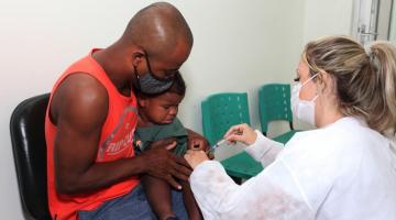 Homem segura uma criança que vai tomar a vacina. Mulher à frente dos dois vai vacinar. #paratodosverem