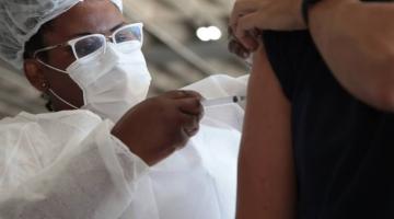 mulher usando máscara e gorro vacina o braço de uma pessoa que segura a manga da blusa. #paratodosverem