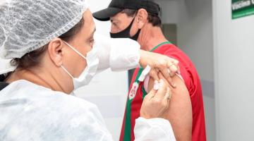mulher usando máscara, gorro e avental vacina homem no braço esquerdo. #paratodosverem