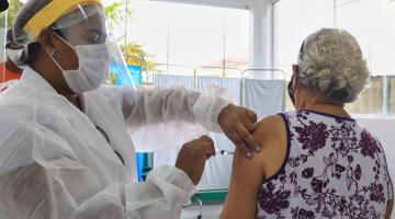 idosa está de costas sendo vacinada no braço esquerdo por uma mulher paramentada com avental, máscara e face shield. #Paratodosverem