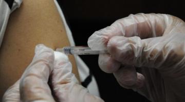 Imagem em close mostra mãos com luvas vacinando um braço. #Paratodosverem