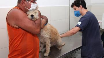 HOmem abraça cachorro que é preparado para receber vacina. Vacinador está usando máscara e aproxima a mão esquerda do animal. #Paratodosverem