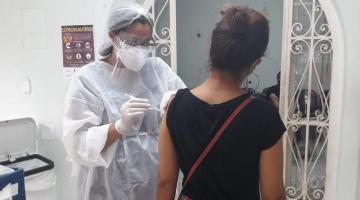Mulher paramentada com avental, luva, gorro e máscaras vacina outra mulher. #Paratodosverem