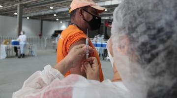 mulher segura seringa com agulha, se preparando para vacina homem que está com a manga da camisa arregaçada. #paratodosverem