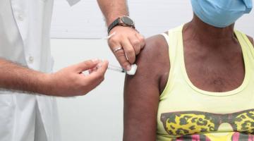 Close de mãos masculinas vacinando no braço direito uma mulher que está sentada. #paratodosverem