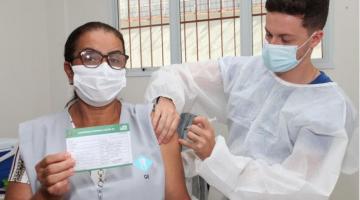 Trabalhadora da saúde mostra carteira de vacinação enquanto recebe vacina no braço por profissional paramentado. #pracegover