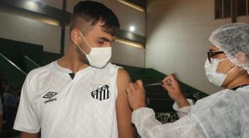 Jovem, com a camisa do Santos, recebe aplicação de vacina no braço por enfermeira paramentada. #pracegover