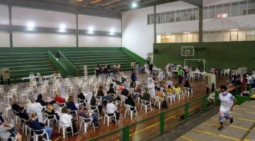 Em um ginásio, várias pessoas sentadas na cadeira aguardando vacinação. #paratodosverem