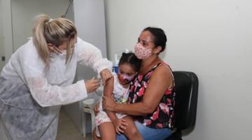 Mulher aplica vacina em criança sentada no colo da mãe #paratodosverem