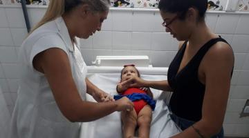 bebê está deitada em maca. Mulher aplica vacina e mãe segura a menina. #paratodosverem
