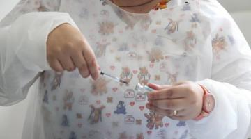 mãos de mulher usando avental estão na frente do tronco inserindo agulha de seringa em frasco de vacina. #paratodosverem